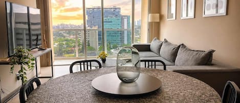 A modern living room with a round table, a sofa, and a large window offering a city view.