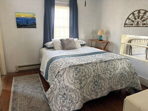 A bedroom with a bed, a nightstand, a lamp, a painting, and a window with curtains. - Riverview Cottage, a quaint downtown home with River and Park views (Millinocket)