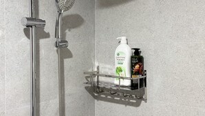 A shower with a handheld showerhead and a wall-mounted faucet. A metal shelf holds two bottles of shampoo and conditioner. - Han House's Apartment 1 (Da Nang)