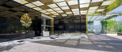 Entrada de hotel con marquesina de vidrio, mostrador de recepción y un logotipo.