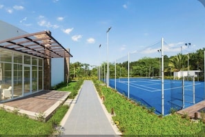Ein Tennisplatz mit Netz, umgeben von Grünflächen und einem Weg, der zu einem Gebäude mit großen Fenstern führt.