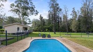 Ein rechteckiger Swimmingpool mit einer gefliesten Liegefläche, umgeben von einem schwarzen Zaun, und ein Haus im Hintergrund.