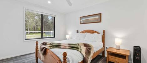 A bedroom with a wooden bed, a nightstand, a desk, a ceiling fan, and a window with a view of trees.