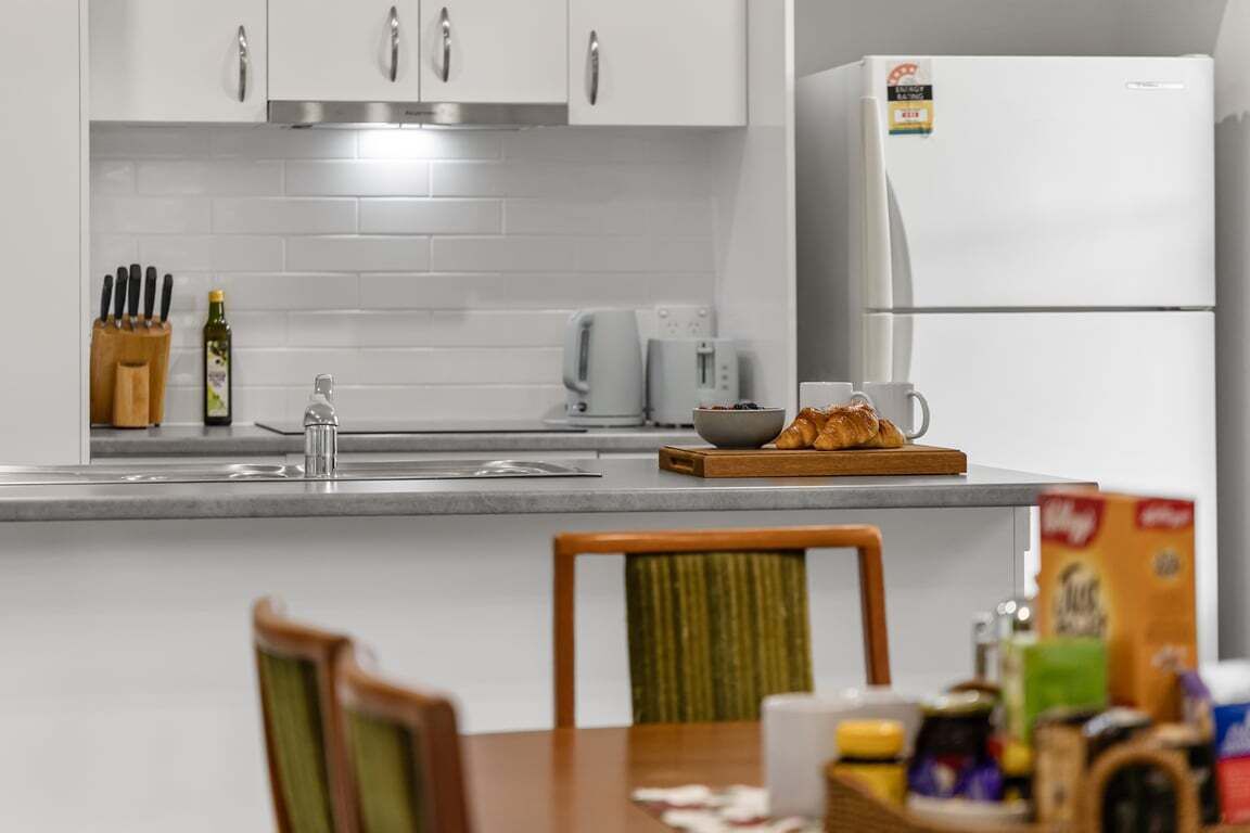 A modern kitchen with white cabinets, a stainless steel range hood, a white refrigerator, and a dining area with a wooden table and chairs.