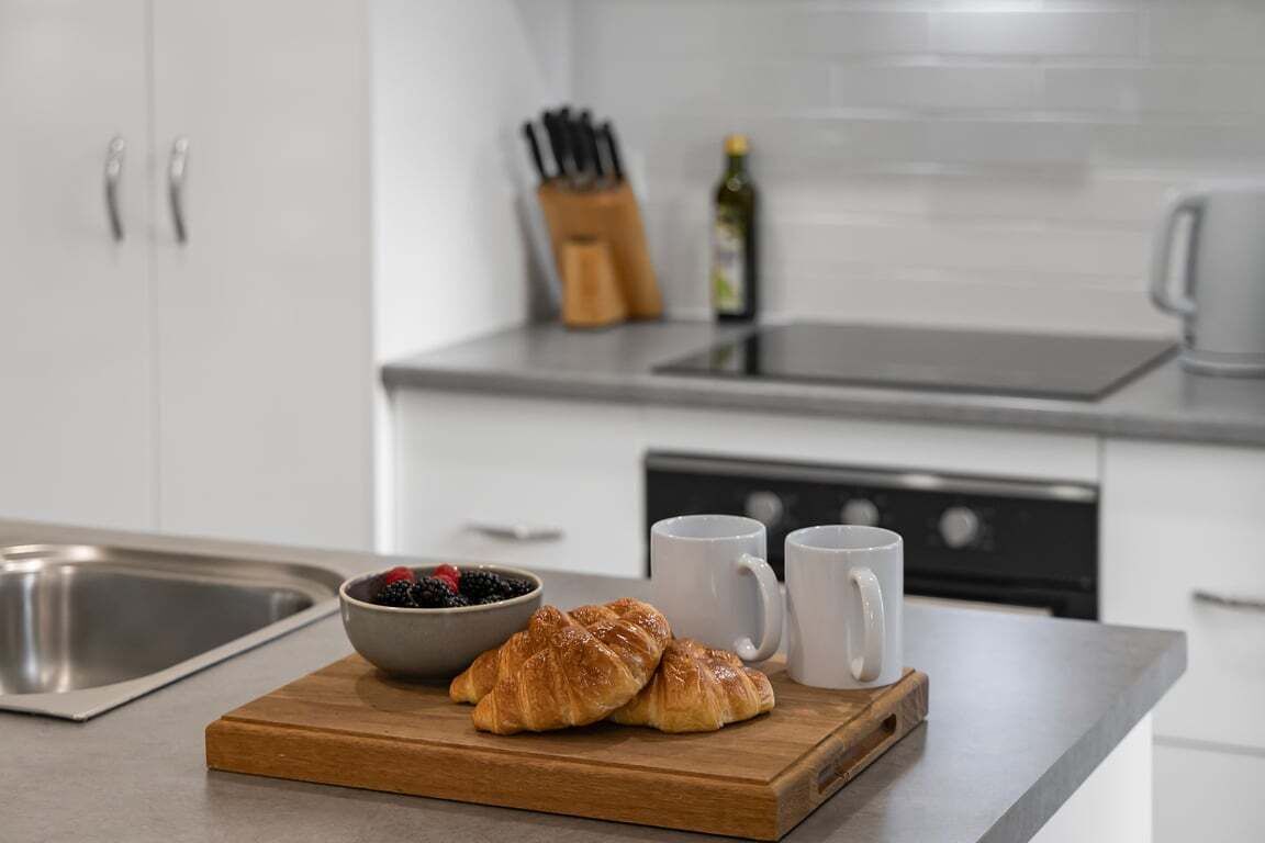 A kitchen counter with a wooden board holding croissants and a bowl of berries, two white mugs, and a knife block in the background.