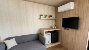 Ein modernes Wohnzimmer mit grauem Sofa, einem fest montierten Flachbildfernseher, einem Holzregal mit Topfpflanzen und einem eingebauten Minikühlschrank.