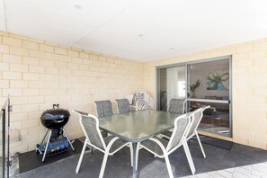 A glass-top table with four chairs, a barbecue grill, and a brick wall. - Spacious 4BR Family Home • Pool with Spa & BBQ (Byford)