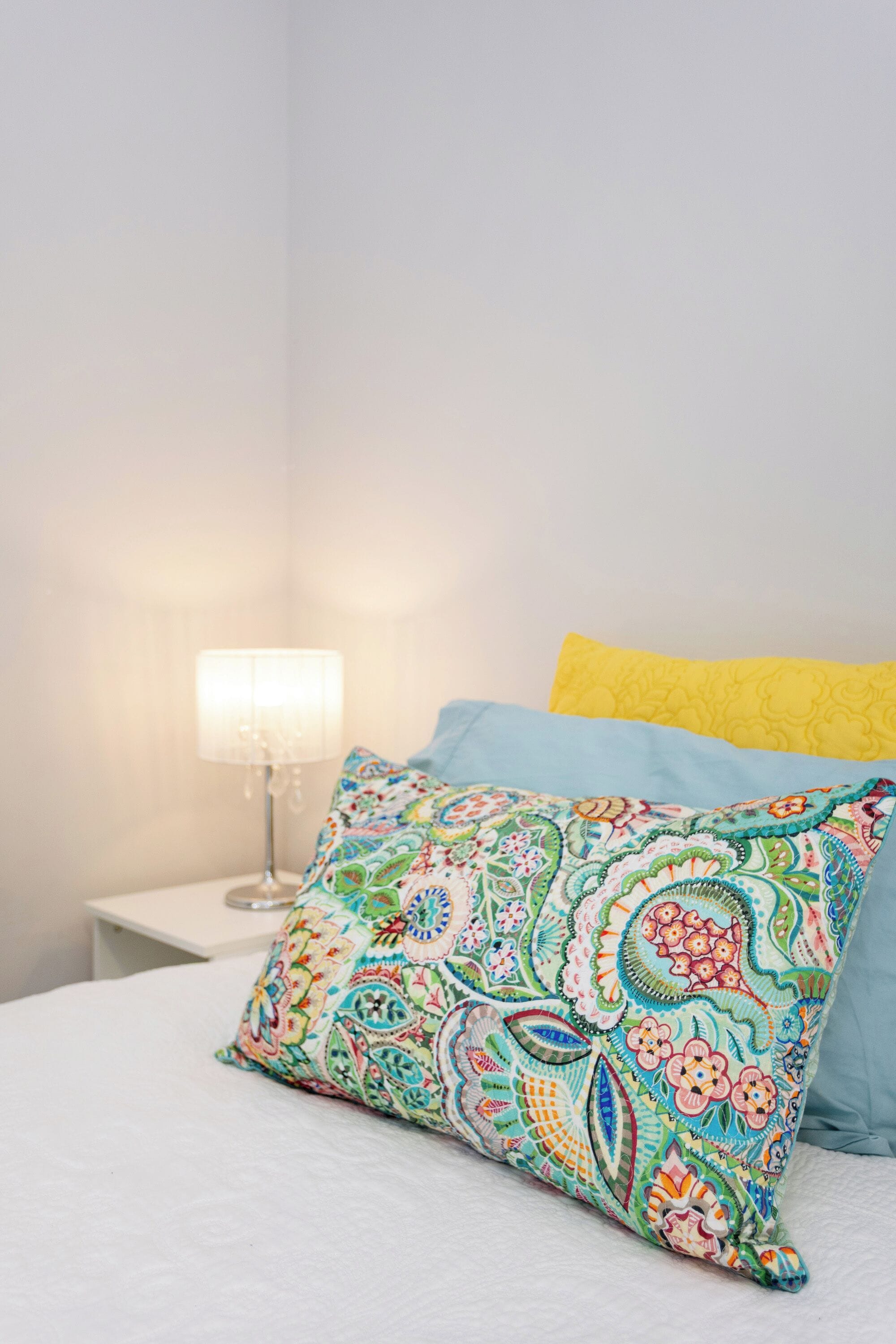 A bed with a colorful patterned pillow and a plain yellow pillow, a white bedside table with a lamp, and a plain wall.