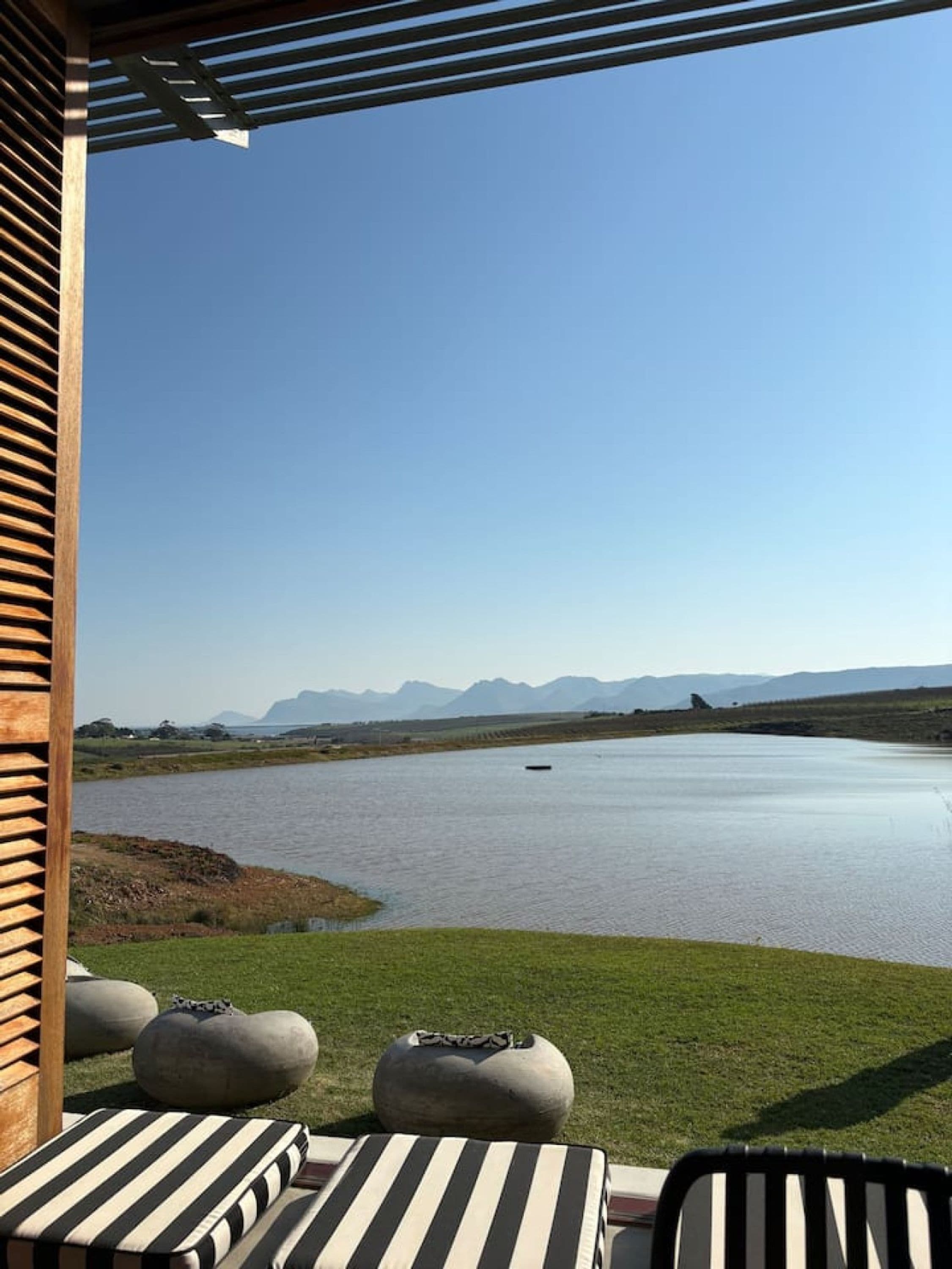 Vista de un lago con montañas al fondo, una pérgola de madera y mobiliario exterior con cojines.