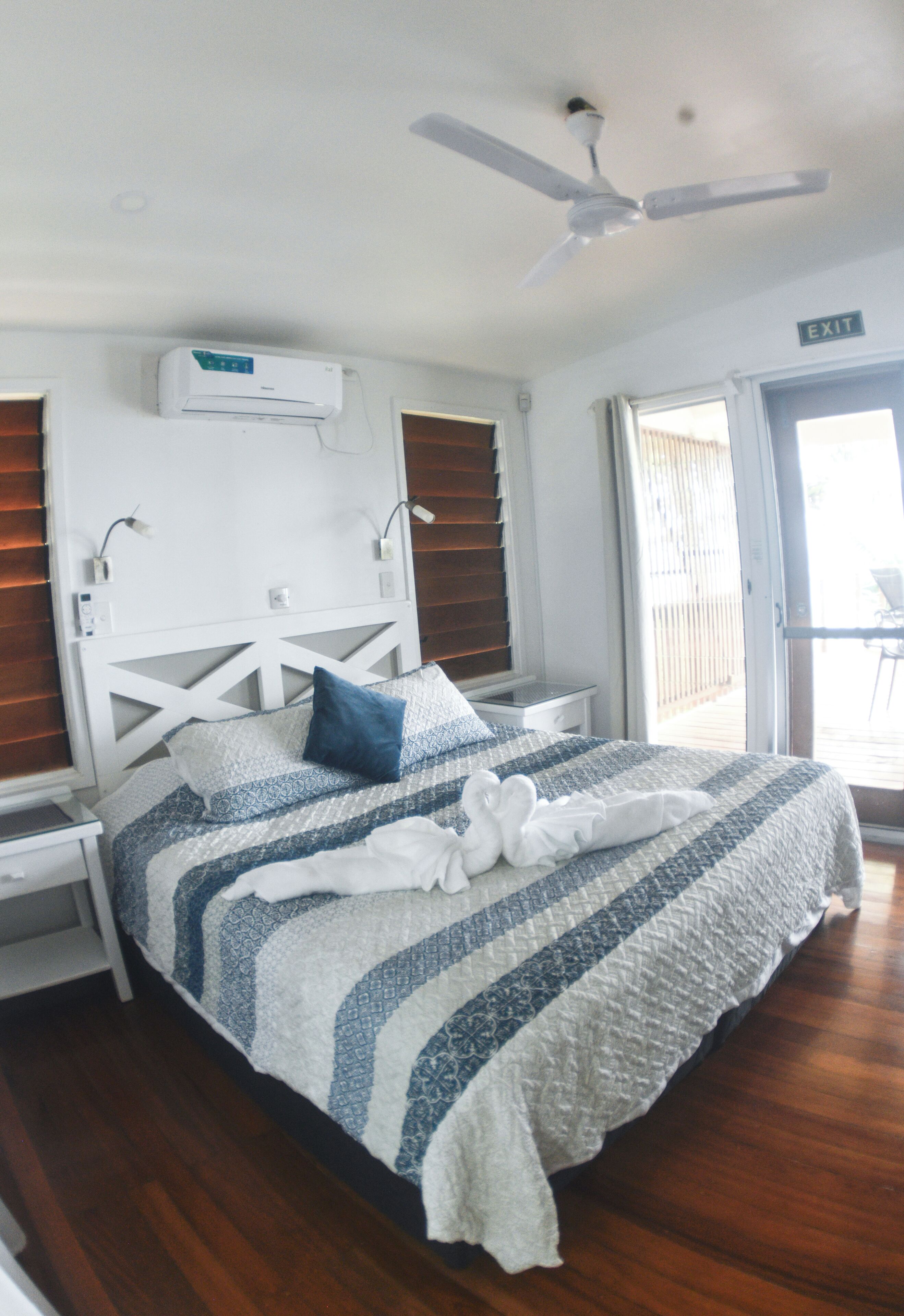 A hotel room with a bed, a ceiling fan, an air conditioning unit, a nightstand, a window with blinds, and an exit sign.