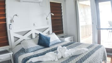 A hotel room with a bed, a ceiling fan, an air conditioning unit, a nightstand, a window with blinds, and an exit sign.