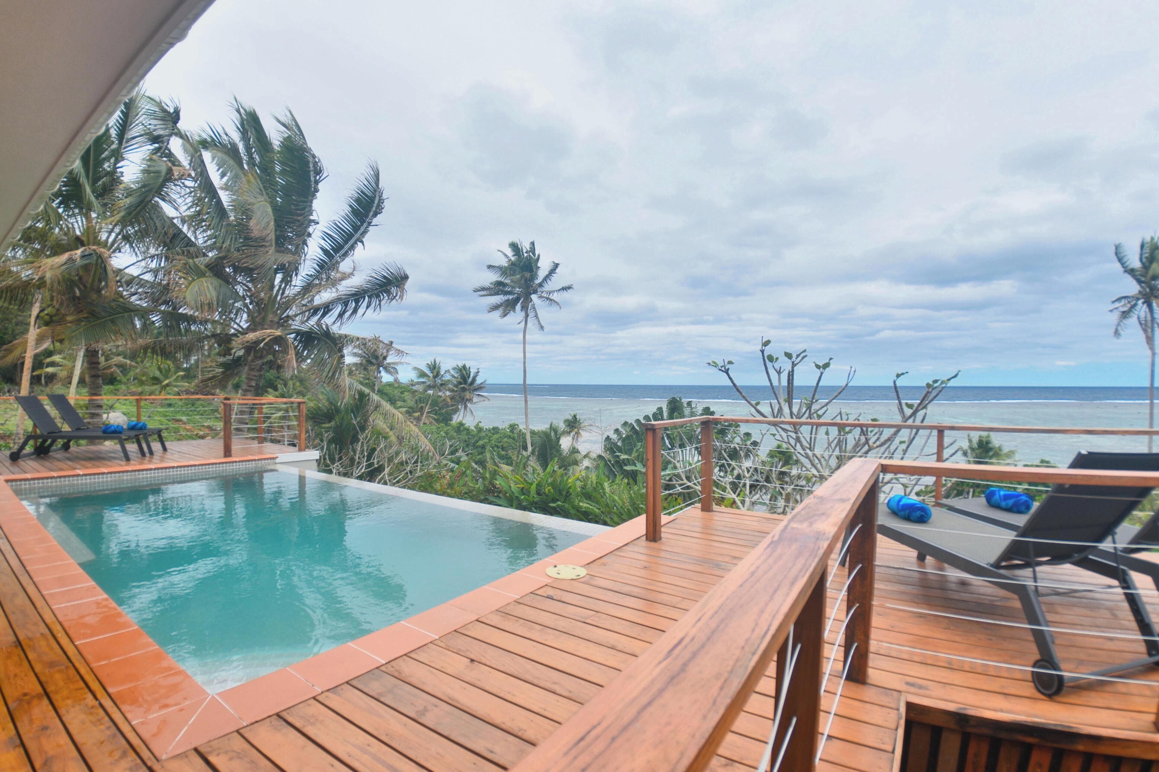 A wooden deck with a pool, lounge chairs, and a view of the ocean and palm trees.