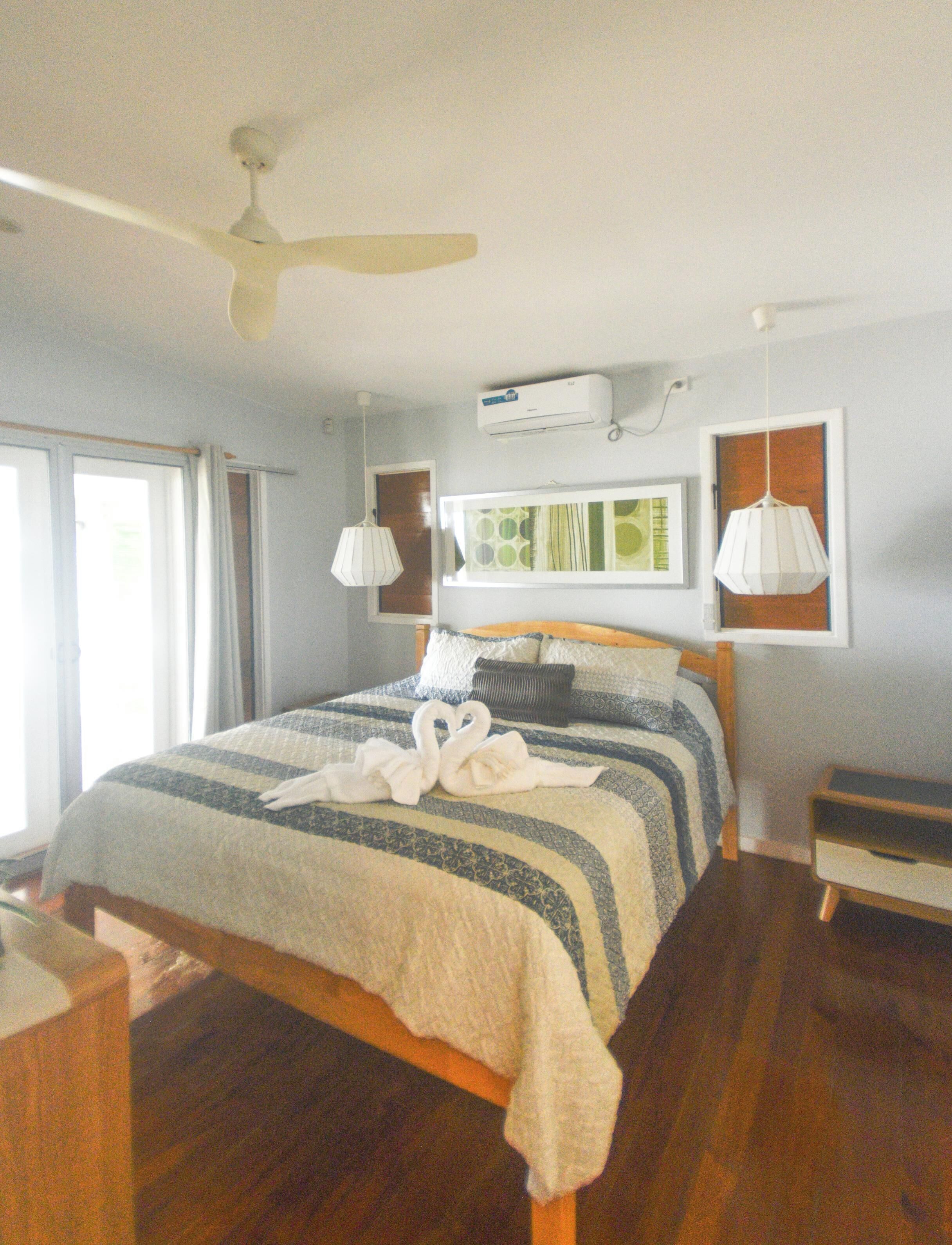 Une chambre d’hôtel comprenant un lit, un ventilateur de plafond, un appareil de climatisation, une fenêtre avec des rideaux et une table de chevet.