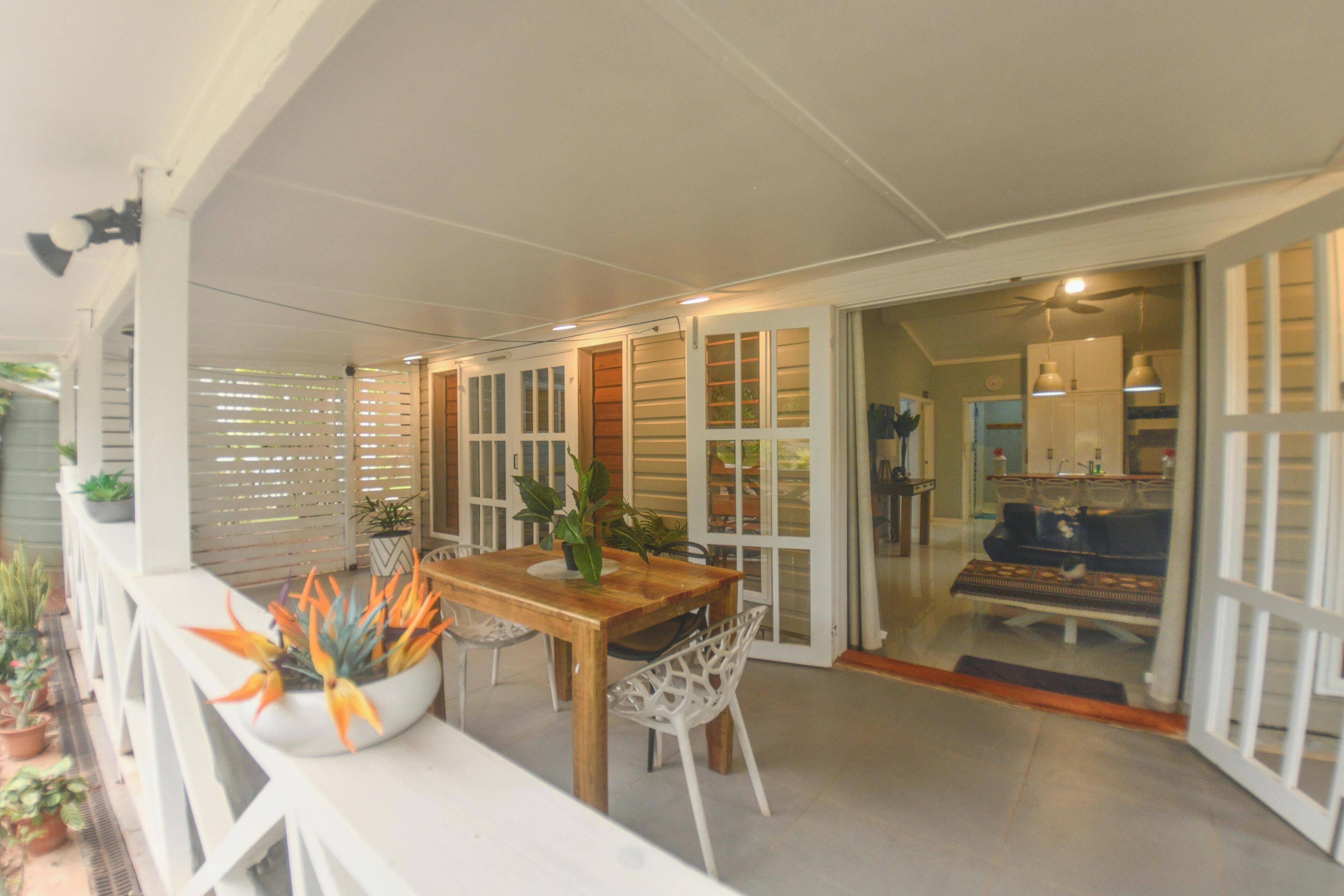 A covered outdoor seating area with a table and chairs, potted plants, and a sliding glass door leading to an interior space.