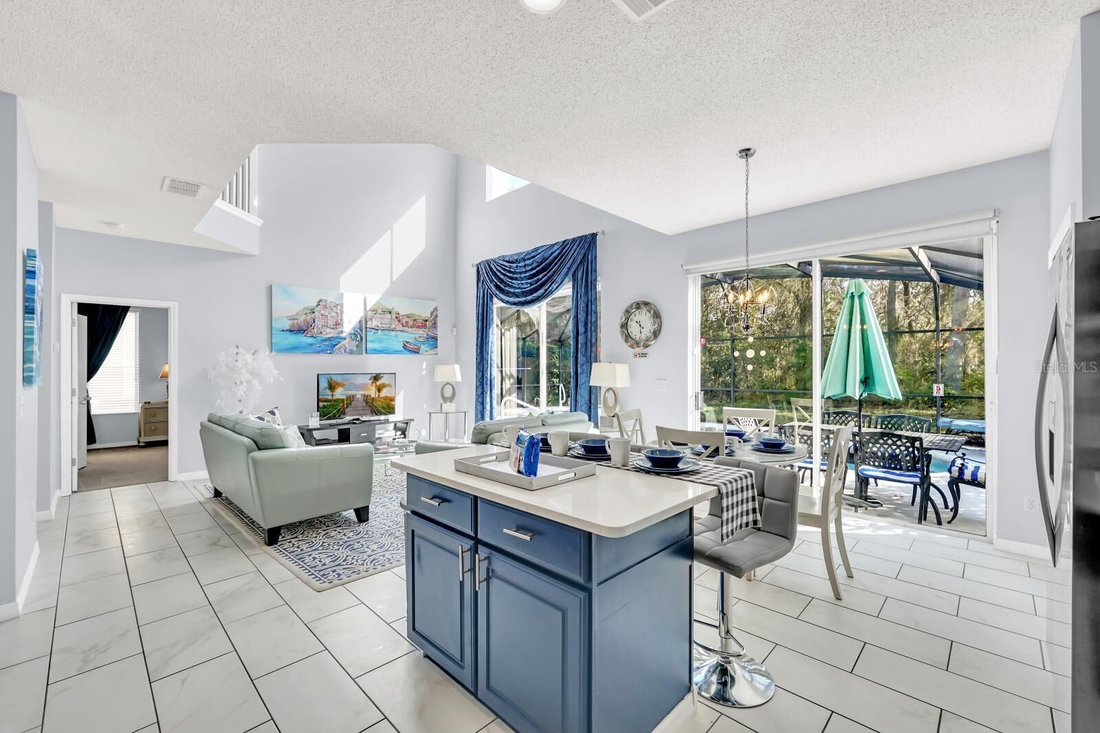 A modern kitchen with a central island, a dining area with outdoor seating, and a living room with a sofa and TV.