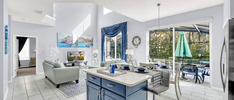 A modern kitchen with a central island, a dining area with outdoor seating, and a living room with a sofa and TV.