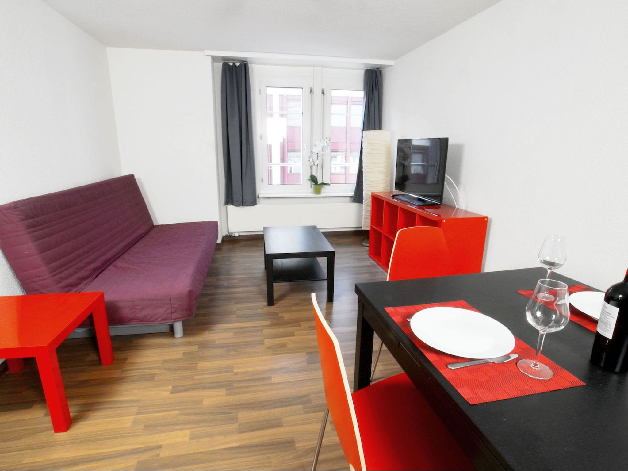 A modern living room with a sofa, a dining table set with plates and wine glasses, a television on a stand, and a window with curtains.