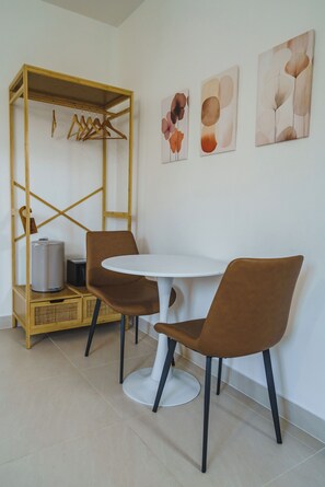 Une pièce comprenant une table ronde, deux chaises marron, un portant en bois où sont suspendus des vêtements, et trois œuvres d’art accrochées au mur.