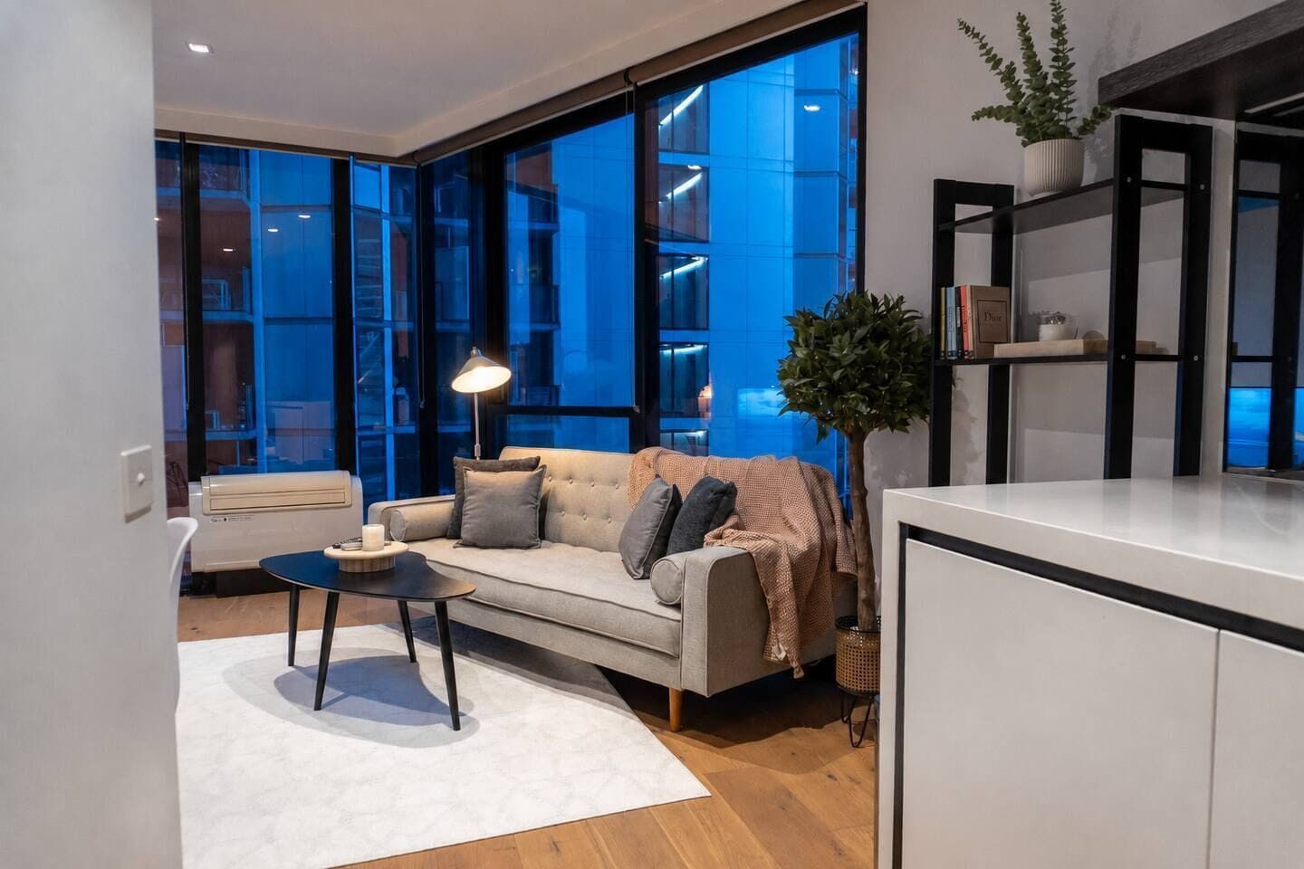 A modern living room with a sofa, a coffee table, and a lamp. There is a large window with a view of a building.
