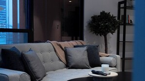 A modern living room with a grey sofa, multiple cushions, a remote control on a side table, and a bookshelf with books and decorative items. - Luxury CBD LaTrobe Retreat 1BR Apt w/Pool & Gym (Docklands)