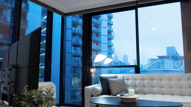 A modern living room with a sofa, coffee table, and a TV. Large windows offer a view of a cityscape and a bridge. - Luxury CBD LaTrobe Retreat 1BR Apt w/Pool & Gym (Docklands)