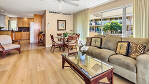 A modern living room with a glass coffee table, a sofa with patterned cushions, wooden dining chairs, and a ceiling fan. - Kaanapali Condo - 1BR/2BA - Beach Cabana, Pools, Private Lanai, A/C (Lahaina)