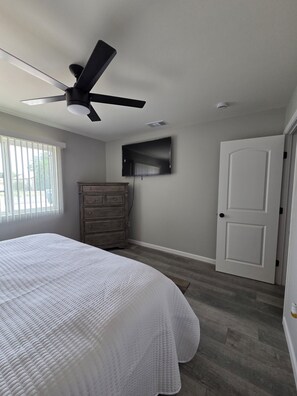 A bedroom with a bed, dresser, ceiling fan, TV, and door.
