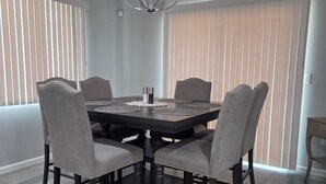 A dining room with a round table, four chairs, a chandelier, and large windows with blinds.