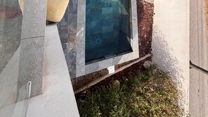 Una piscina con borde de piedra, un pequeño solárium con sillas y una estructura de madera al fondo.