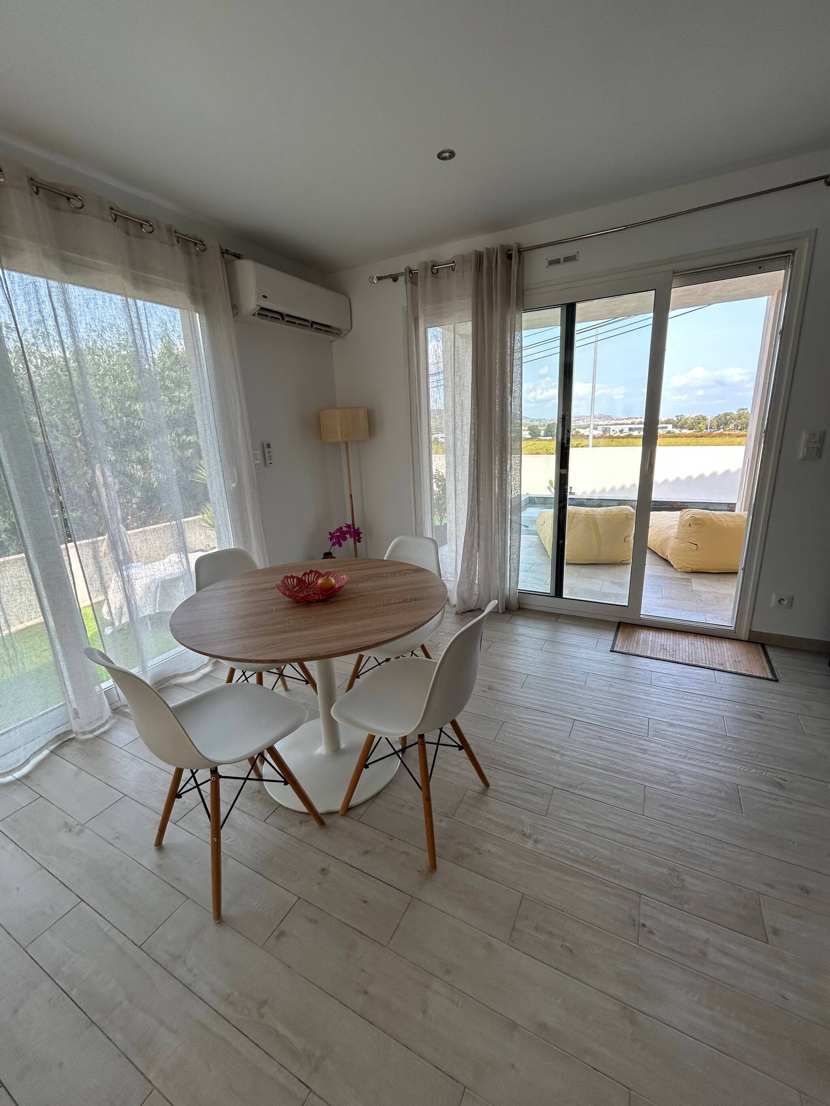 A dining area with a round table, white chairs, and a view through large glass doors.