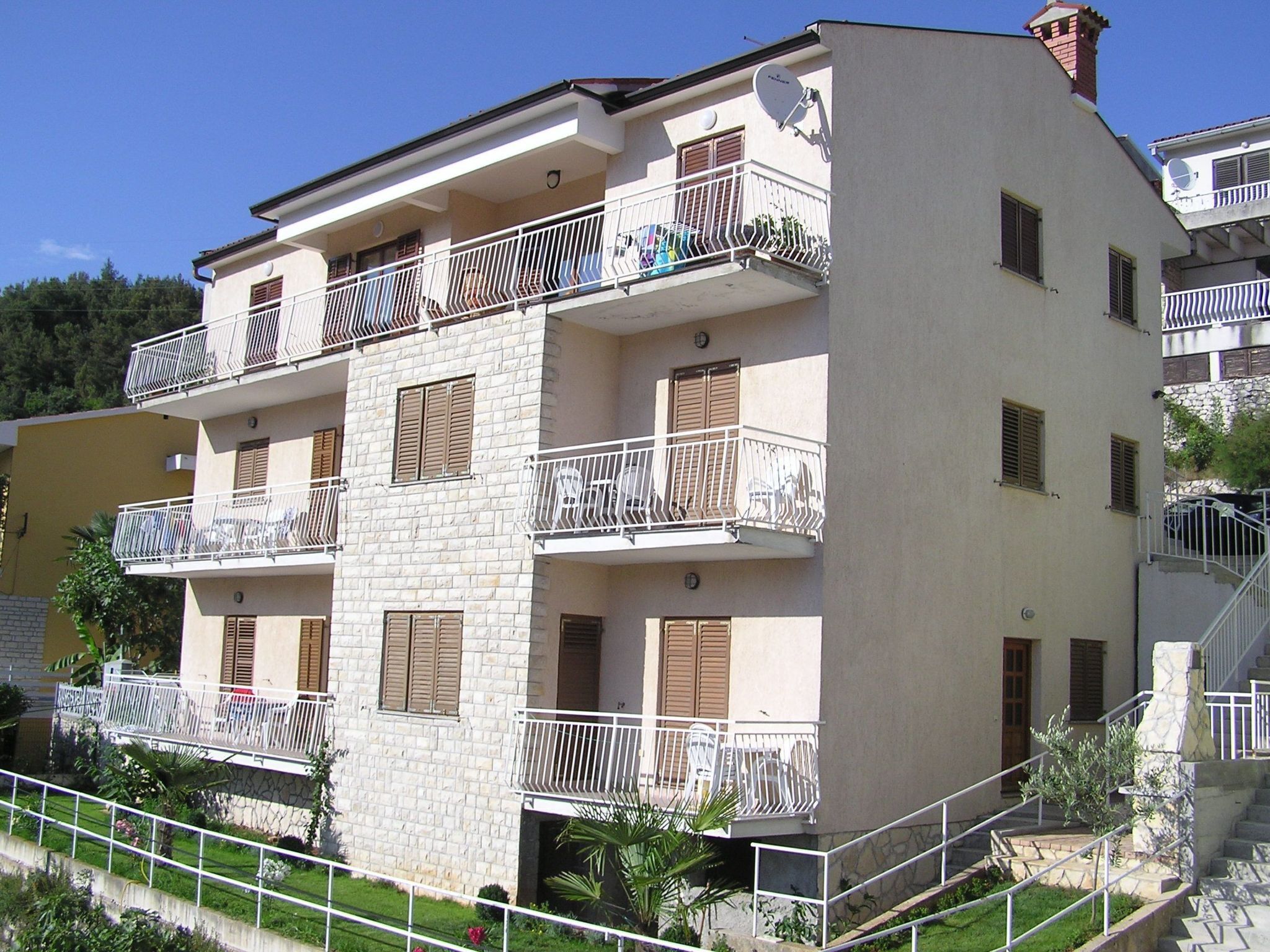 A multi-story residential building with balconies, a stone facade, and a staircase leading to the entrance.