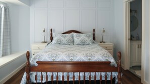 Une chambre à coucher avec un lit en bois, des murs blancs, une fenêtre avec un rideau et une porte donnant sur une salle de bain.