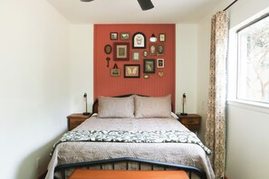A bedroom with a bed, bedside tables, a wall with framed pictures, and a window with curtains. - Texan Cottage Getaway —Near UTSA, Six Flags, Seaworld; Family Recreation & Trail (San Antonio)