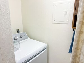 Ein kleiner Waschraum mit einer weißen Waschmaschine, einer fest installierten Steckdose an der Wand und einer Tür mit Lichtschalter.
