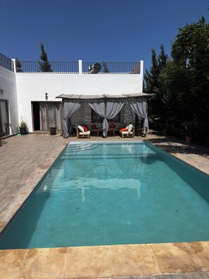 A swimming pool with a covered seating area and a white building in the background. - Villa Sylvester, Essaouira (Essaouira)