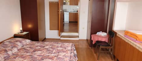 A bedroom with a bed, a desk, a chair, a wardrobe, and an open door leading to a kitchen.