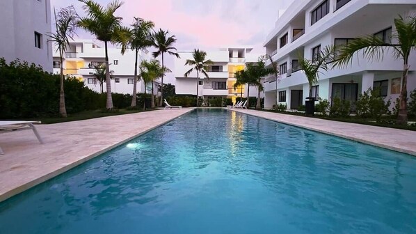 Ein Pool mit Liegestühlen, umgeben von Palmen und modernen Gebäuden.