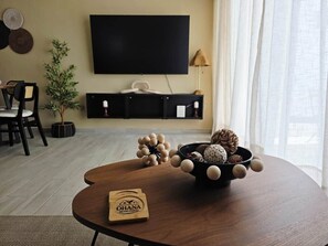 Un salon moderne avec une table basse ronde en bois, un bol noir contenant des objets décoratifs, un téléviseur fixé au mur et un coin repas avec des chaises et une petite plante en pot.