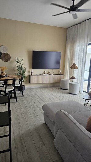 Ein modernes Wohnzimmer mit einem Flachbildfernseher, einem Deckenventilator, einer Essecke mit Tisch und Stühlen sowie einem großen Fenster mit Vorhängen.