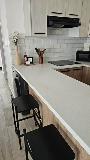 Eine moderne Küche mit weißer Arbeitsplatte, Holzschränken und einem schwarzen Herd. Auf der Arbeitsplatte stehen zwei schwarze Barhocker und eine Vase mit Blumen.