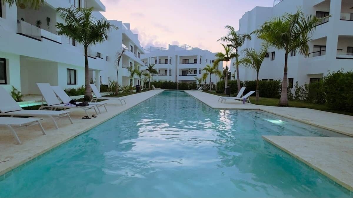 Una piscina con sillones de descanso, rodeada de edificios blancos y palmeras.