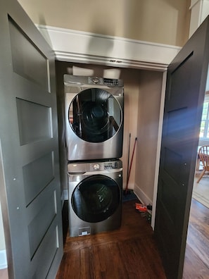 A compact laundry room with two stacked front-loading washing machines, a broom, and a red box on the floor.