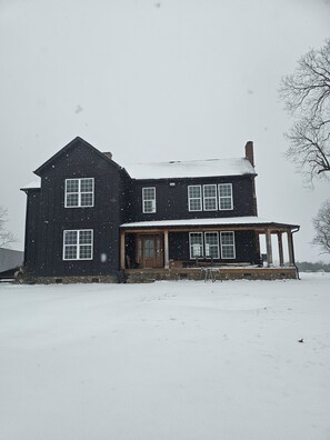 A large, two-story wooden house with a porch and multiple windows, surrounded by snow.