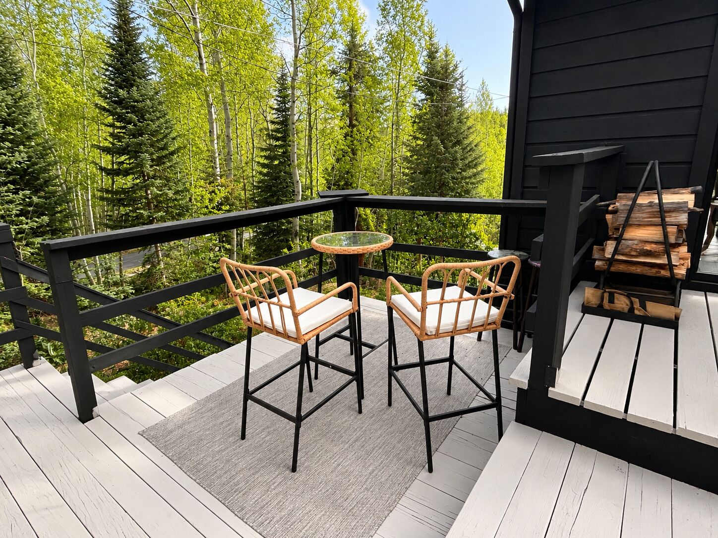A deck with two wicker chairs and a small table, surrounded by a black railing, overlooking a forested area.