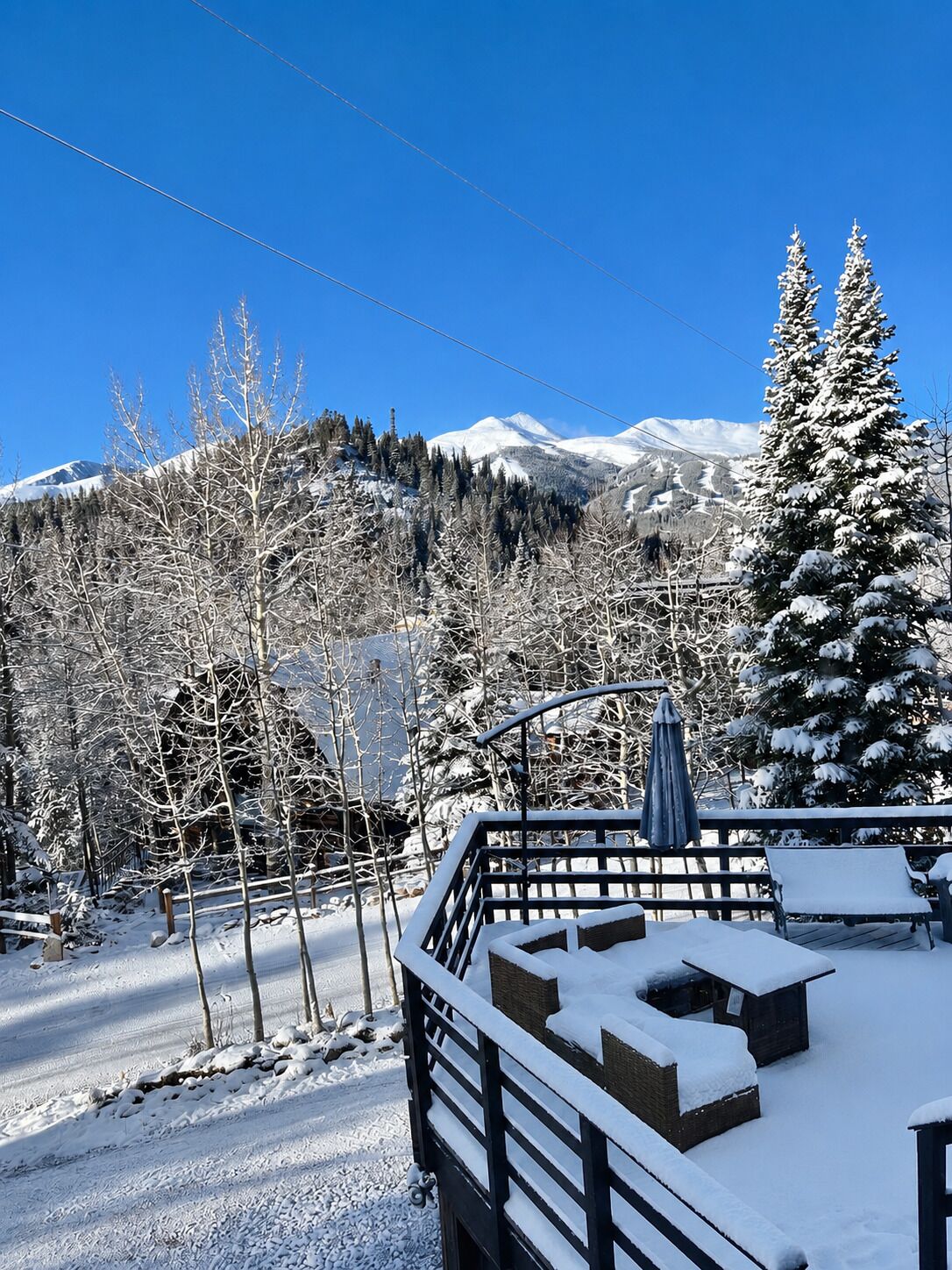 A snow-covered deck with outdoor seating, a view of snow-covered trees, and a mountain in the background.