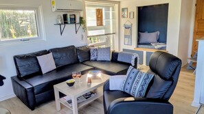 A modern living room with a black leather sofa, a coffee table, and a chair. There are two windows with blinds, a shelf with books and a plant, and a bed with a blue headboard. - Cozy holiday home near the beach (Haderslev)