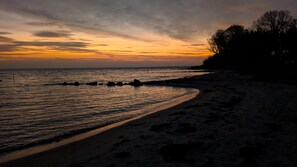 Ein Strand mit einer geschwungenen Küstenlinie, ruhigem Wasser und einem Sonnenuntergangshimmel.