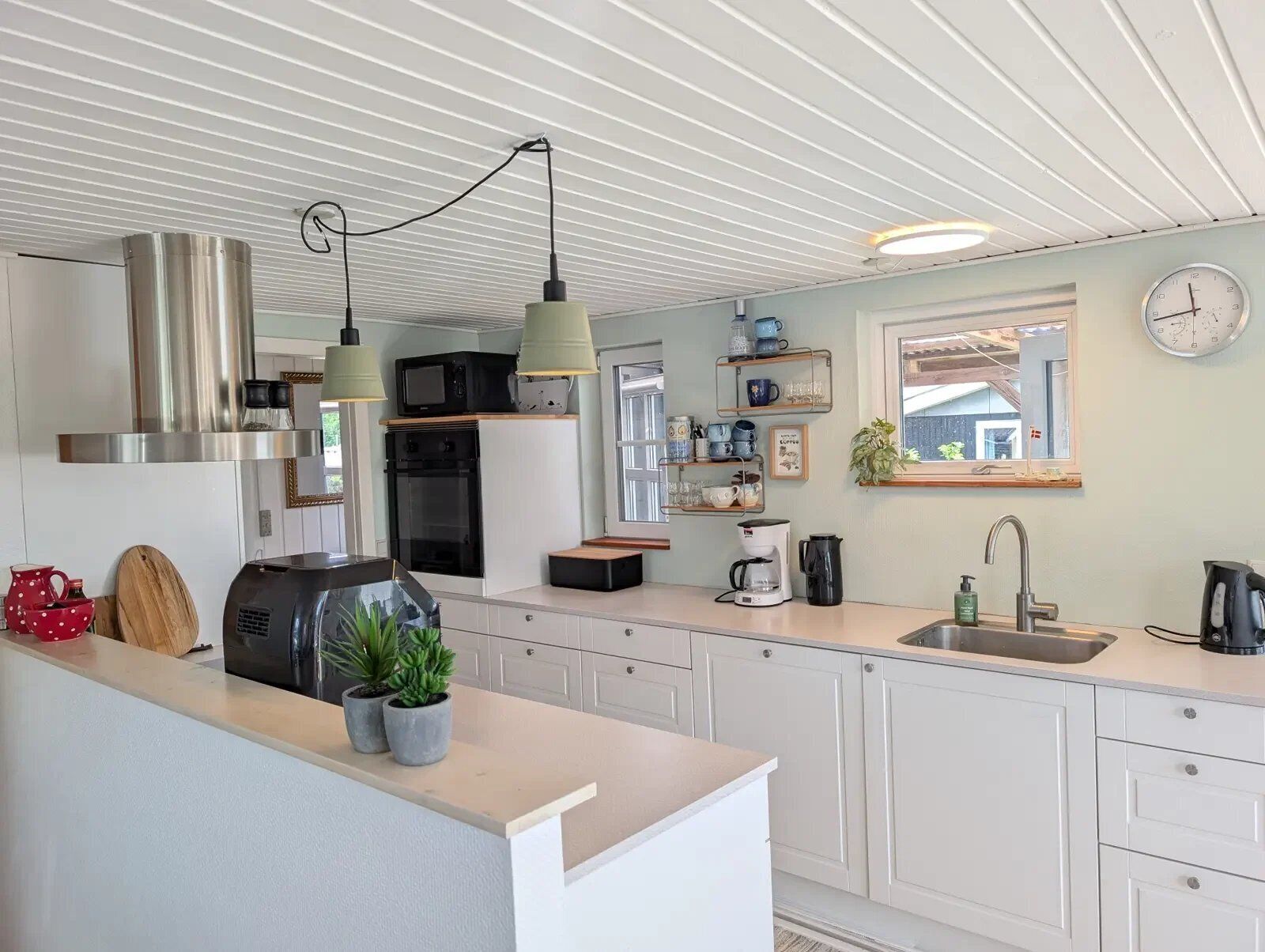 Una cocina moderna con armarios blancos, campana extractora de acero inoxidable, luces colgantes, cafetera y fregadero.