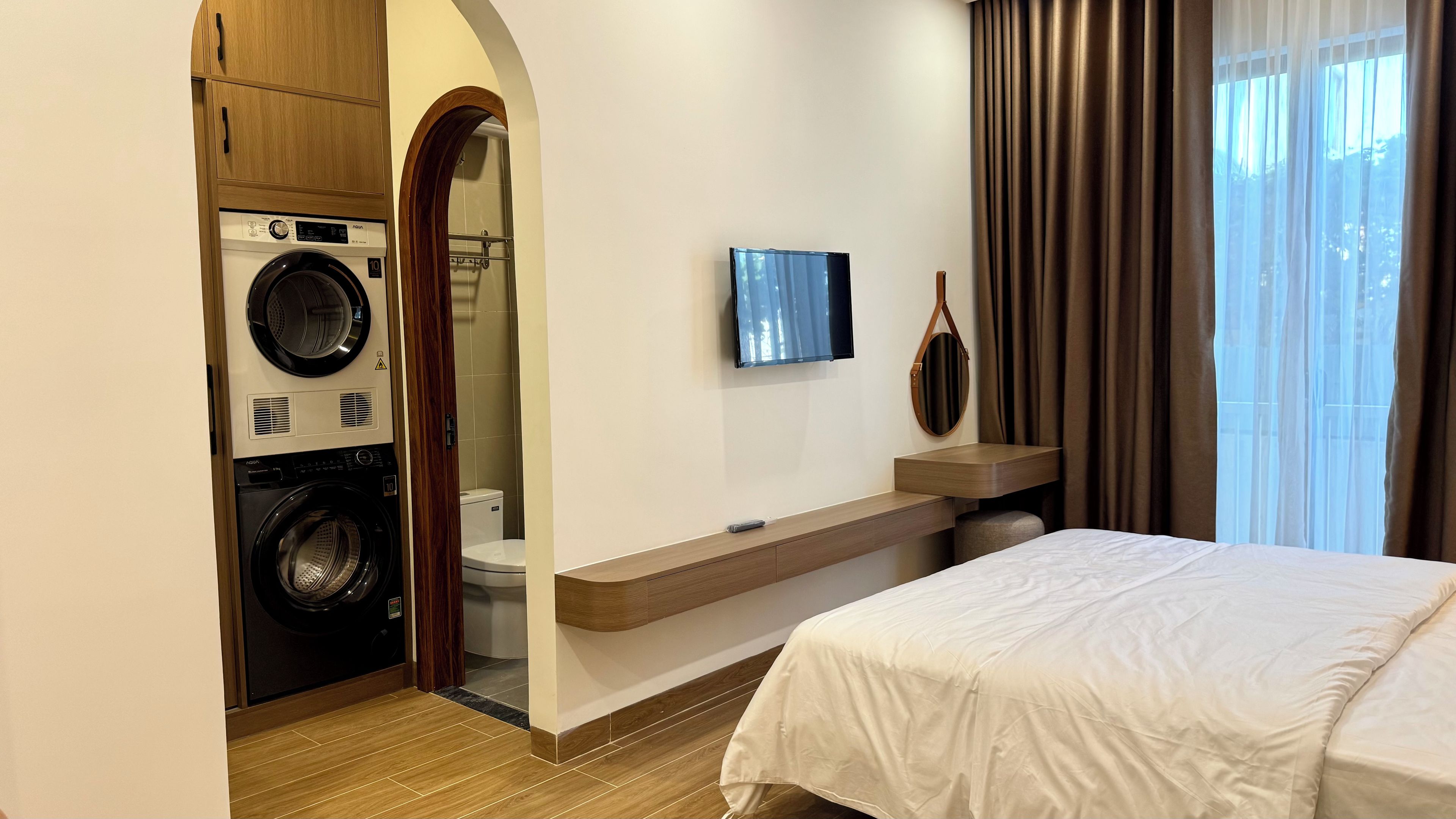 A hotel room with a bed, a TV, a mirror, a small table, a washing machine, and a toilet.