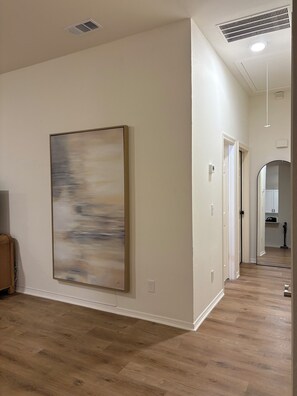A hallway with a large abstract painting, wooden flooring, and a door leading to a kitchen area. - Spacious 3BR House in Frisco - Minutes from Top Event Attractions (Frisco)